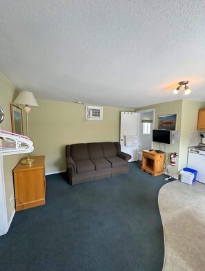 A living room with a sofa, a lamp, a television, a small cabinet, and a kitchenette area.