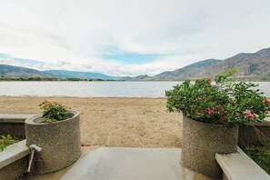 A beach with a concrete pathway leading to a seating area, a body of water, and mountains in the background.
