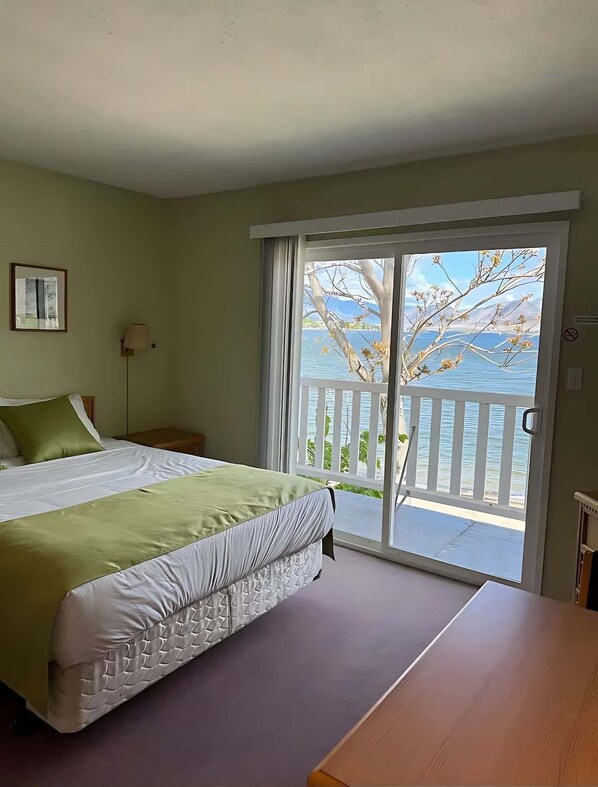 A bedroom with a bed, a nightstand, a lamp, a view of a balcony with a railing, and a door.