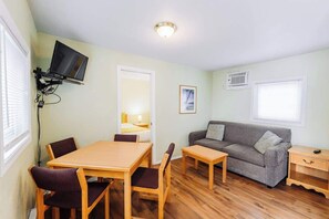A room with a dining table, chairs, a sofa, a TV mounted on the wall, a bedside table, and a window with blinds.