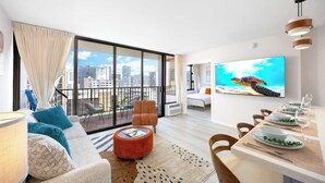 A modern living room with a large window, a sofa, a dining table set, and a TV displaying a sea turtle. - 14Fl Renovated Ocean View with Pool & Parking (Honolulu)
