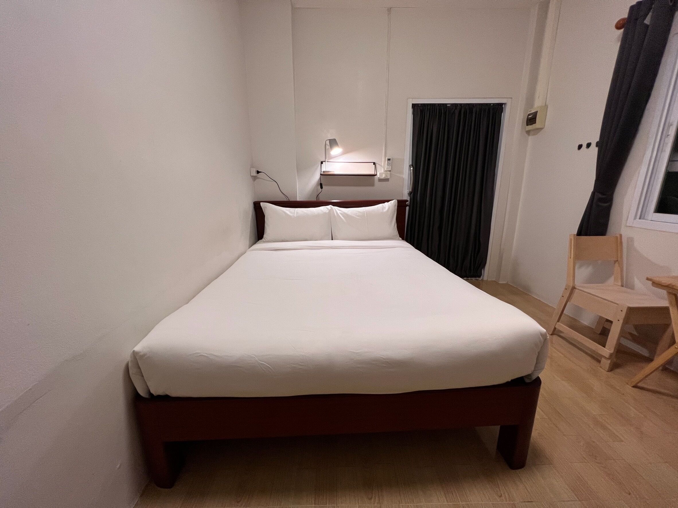 Una cama individual con estructura de madera, una mesita de noche con lámpara, una silla y una ventana con cortinas.