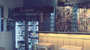 A bar area with two refrigerators stocked with beverages, a wall of bottled drinks, and two stools. - Naisang Hostel (Chumphon)
