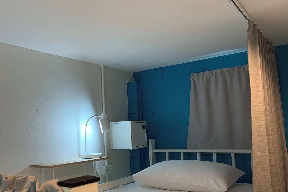 Una cama individual con ropa blanca, una mesita de noche con lámpara, pared azul y cortina.