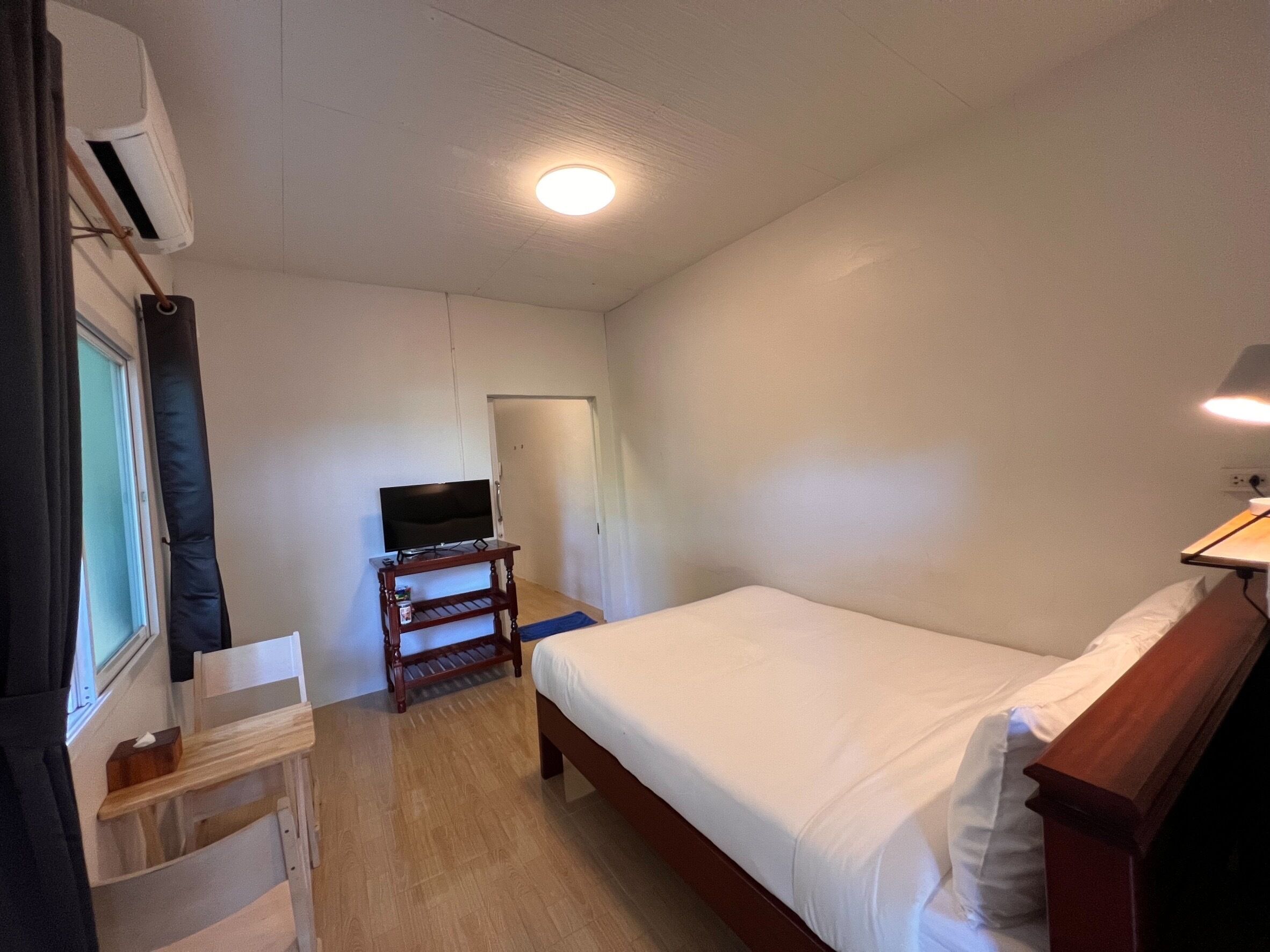 Een slaapkamer met een bed, een televisie op een standaard, een bureau met een stoel en een raam met gordijnen.