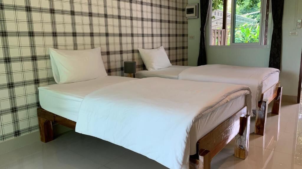 A room with two beds, a checkered wall, a window with a view, and a small table with a lamp.