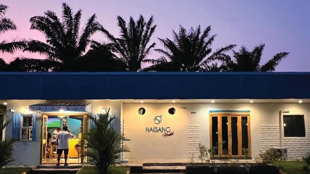 A hotel entrance with a sign reading 'NAISANG Hotel,' palm trees in the background, and a person standing at the entrance. - Naisang Hostel (Chumphon)