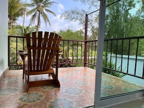 A wooden chair on a balcony with a tiled floor, a railing, and a view of a body of water and palm trees.