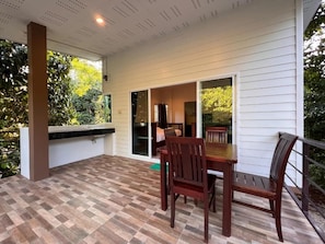 An outdoor patio with a wooden table and chairs, a white exterior wall, and a view of greenery.