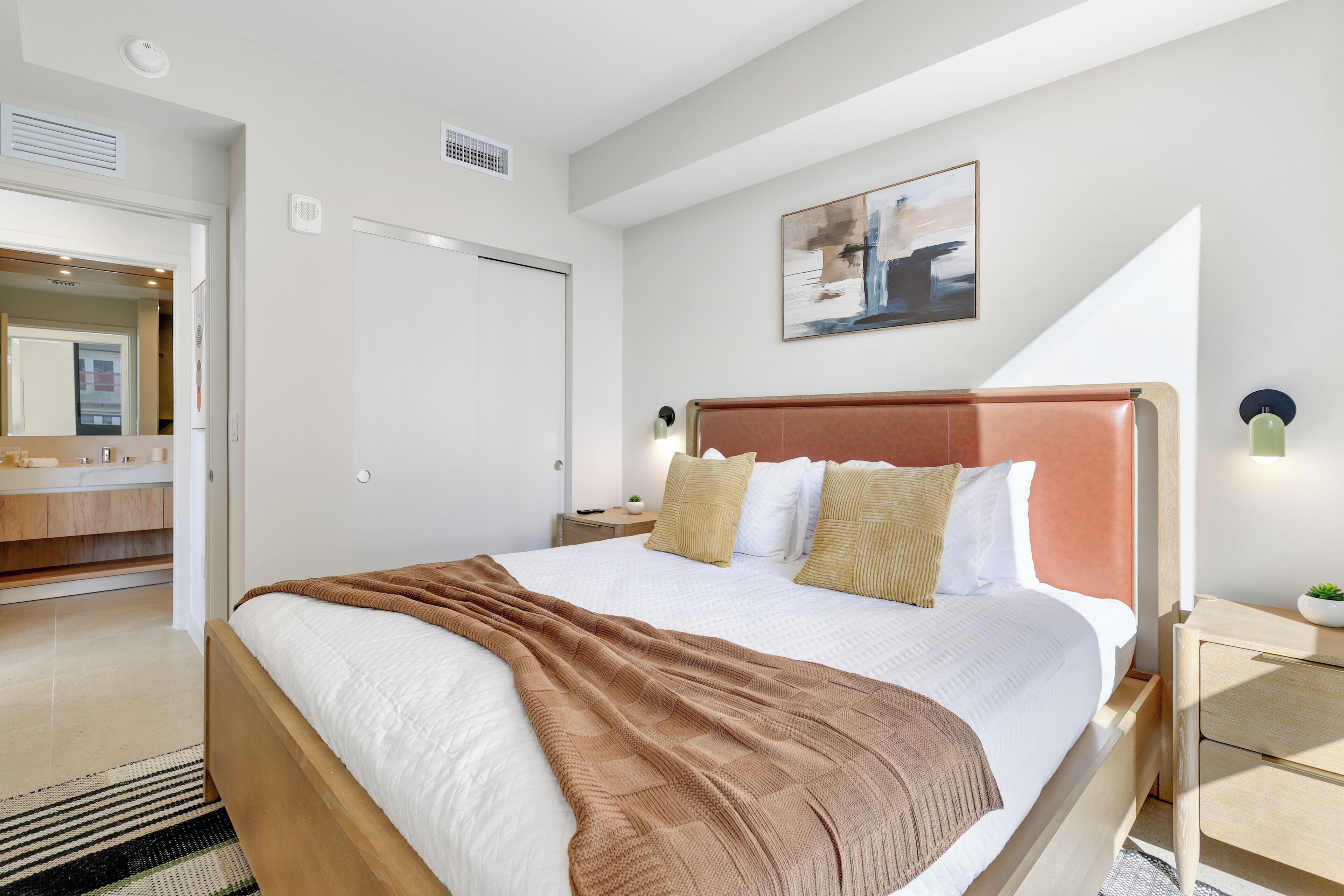 Ein modernes Hotelzimmer mit einem großen Bett, einem Nachttisch mit Lampe, einer kleinen Pflanze und einem Bild an der Wand.