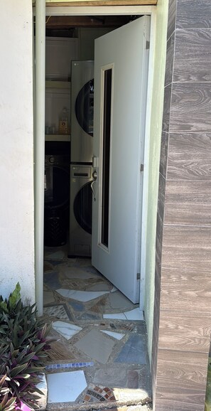 Une porte donne accès à une buanderie équipée d’un lave-linge et d’un sèche-linge, avec un sol carrelé et une plante en pot.