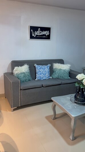 A grey sofa with decorative pillows, a 'Welcome' sign, and a small table with a vase. - Fresh country air, tranquility, pool, and BBQ—pets welcome. (Bonao)