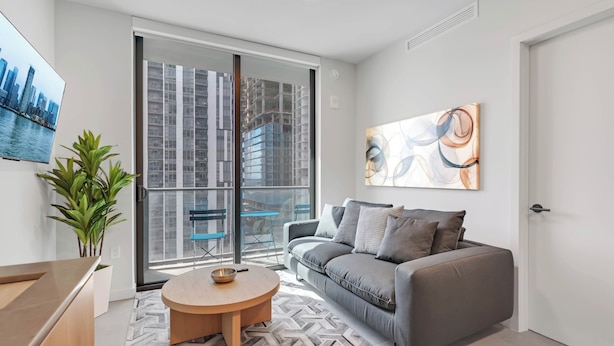 A modern living room with a grey sofa, a round wooden coffee table, a potted plant, and a large window with a city view. - AEA - 121 NE 5th St Miami (Miami)