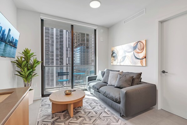 A modern living room with a grey sofa, a round wooden coffee table, a potted plant, and a large window with a city view. - AEA - 121 NE 5th St Miami (Miami)
