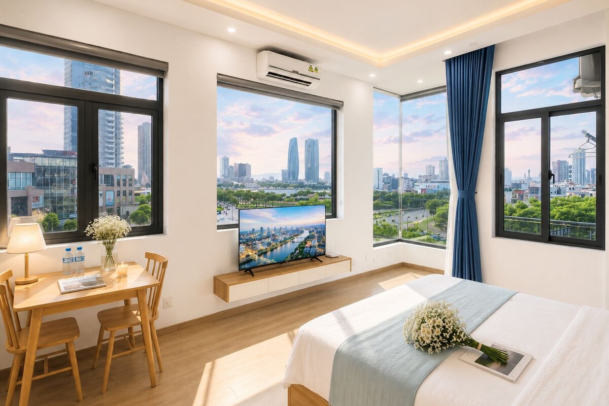 A hotel room with a bed, a desk with a chair, a TV mounted on the wall, and large windows offering city views.