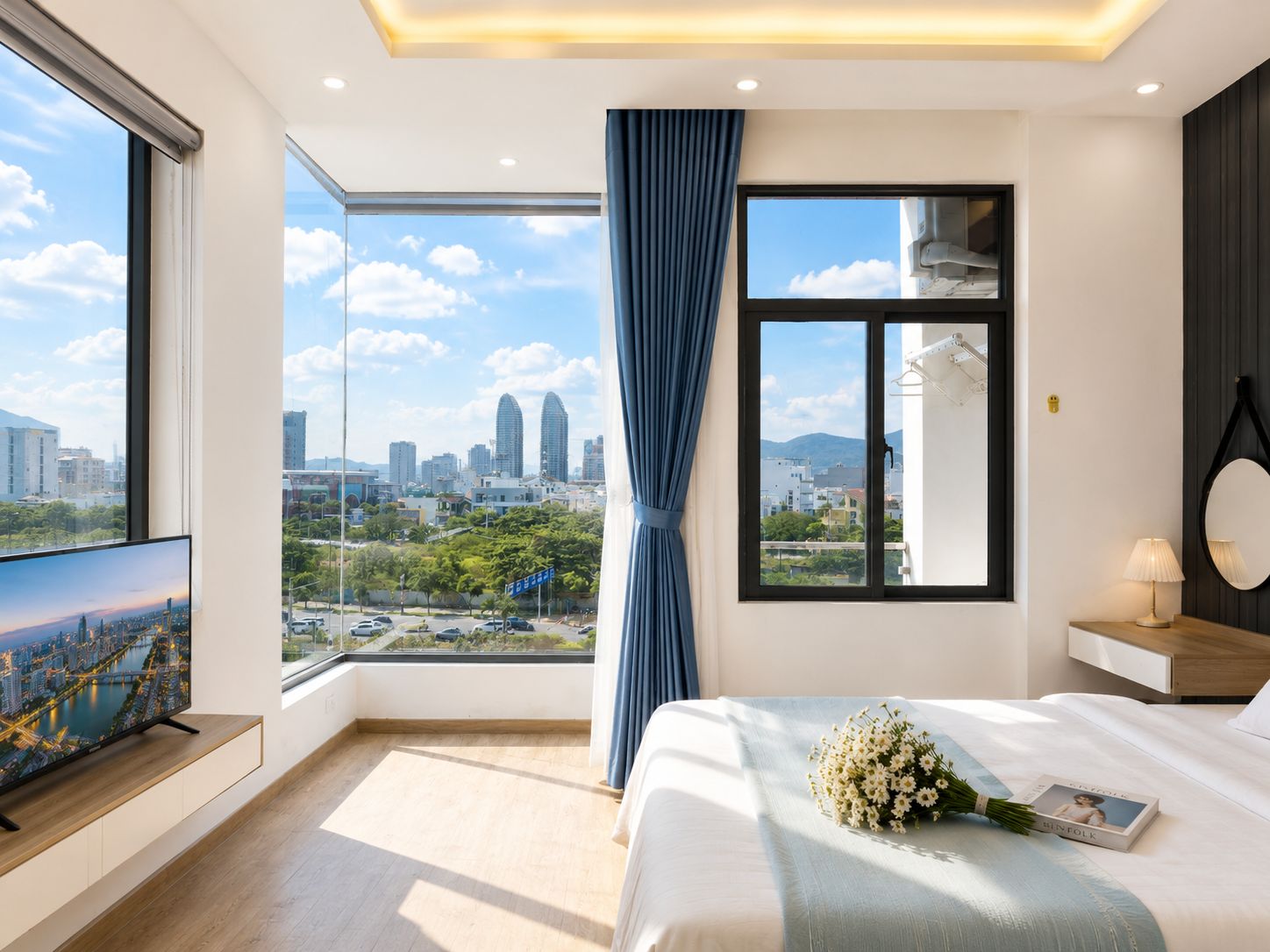 A modern hotel room with a large window, a flat-screen TV, a bed with a bouquet of flowers, and a bedside table with a lamp.
