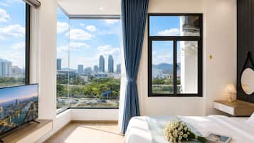 A modern hotel room with a large window, a flat-screen TV, a bed with a bouquet of flowers, and a bedside table with a lamp.