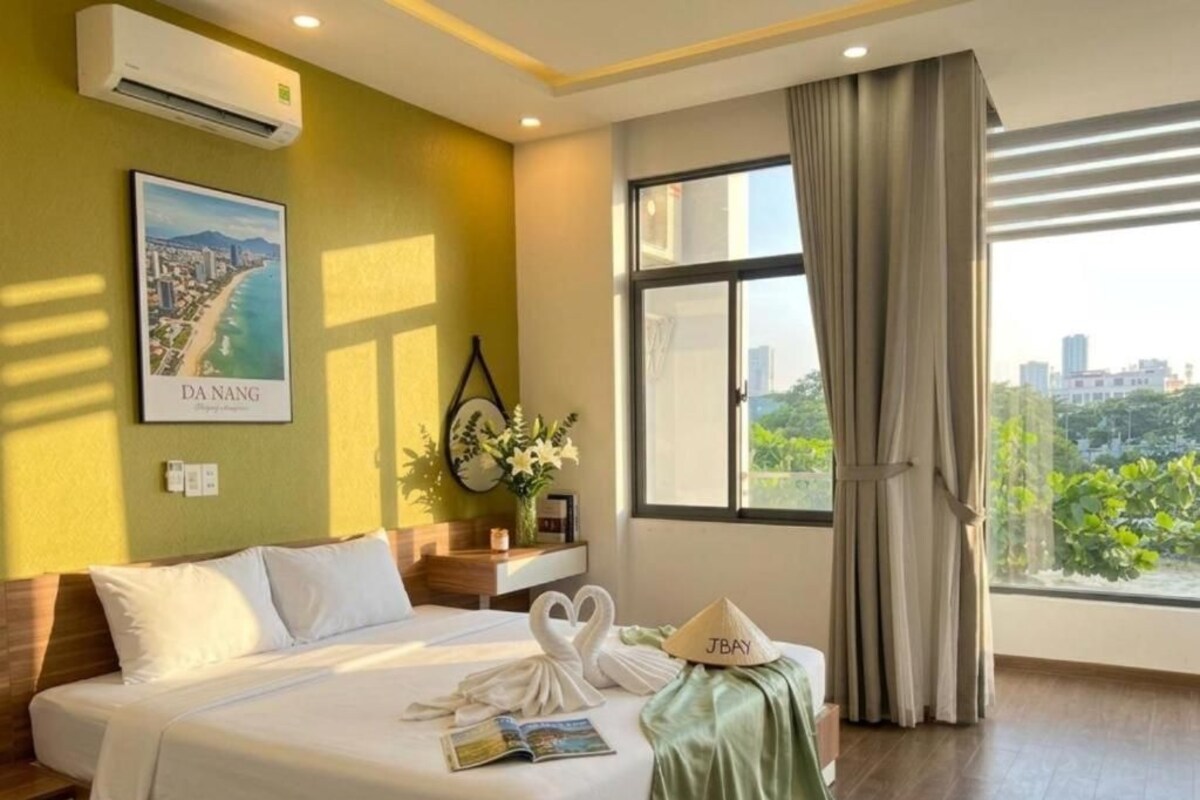 A hotel room with a bed, a window with curtains, a wall-mounted air conditioner, a framed picture, and a bedside table with a vase of flowers.