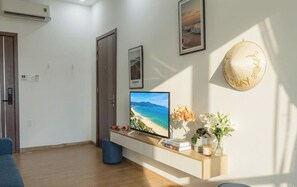 A hotel room with a large TV displaying a beach scene, a wooden shelf with books and a vase of flowers, and a decorative hat on the wall. - JBAY Riverfront Boutique Hotel (Da Nang)