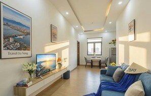 A modern hotel room with a large TV, a sofa, a dining area, and a cityscape view. - JBAY Riverfront Boutique Hotel (Da Nang)