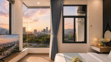 A modern hotel room with a large bed, a television, a window with a city view, and a bedside table with a lamp and flowers.