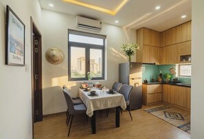 A dining area with a table set for four, a window with a city view, and a kitchen with wooden cabinets. - JBAY Riverfront Boutique Hotel (Da Nang)
