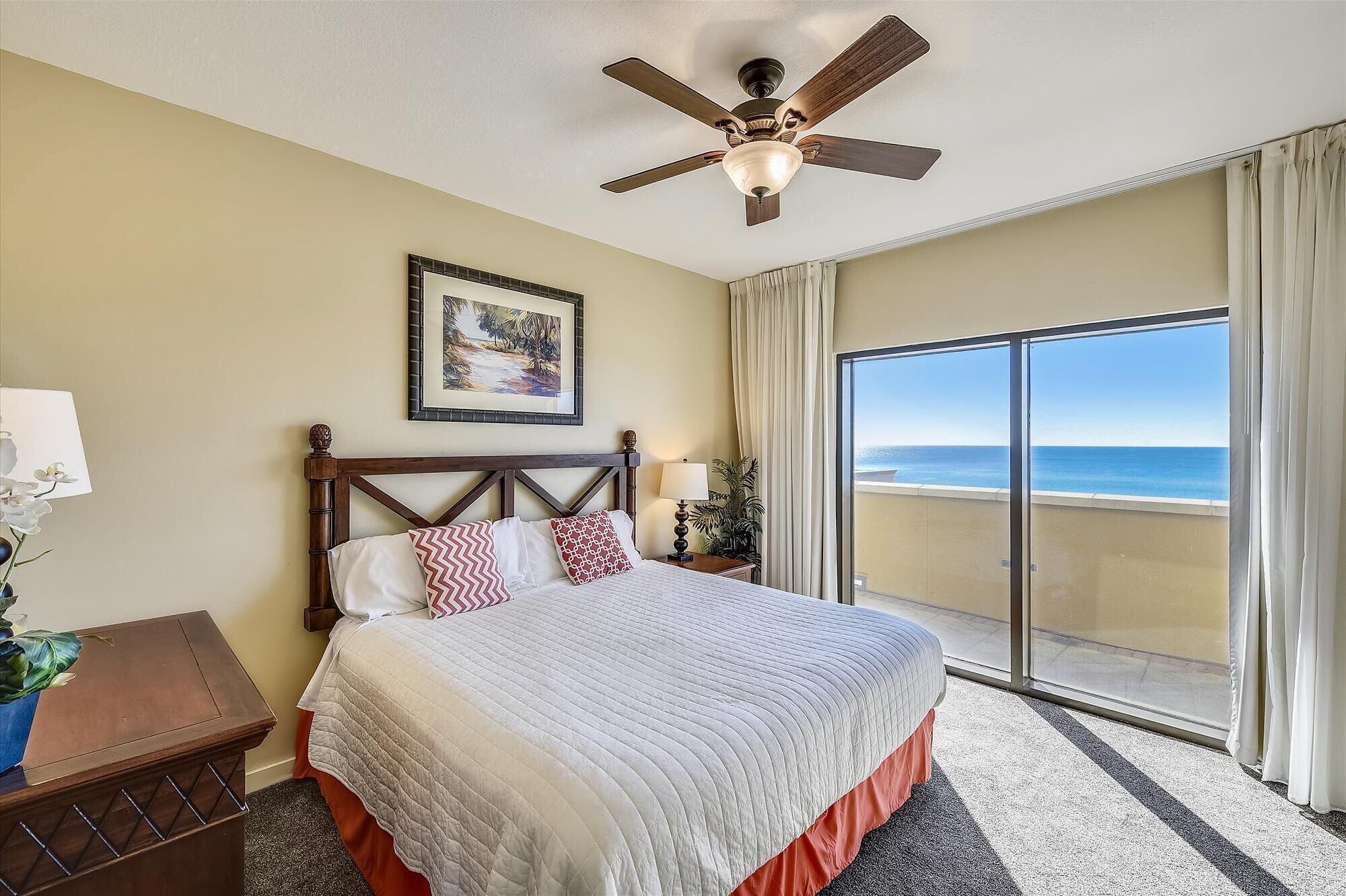 A hotel room with a bed, a ceiling fan, a painting, a nightstand, a lamp, and a view of the beach through a sliding glass door.