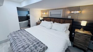 Ein modernes Hotelzimmer mit einem großen Bett, einem Nachttisch mit Lampe, einem Holzkopfstück, einer eingebauten Bank und einem im Hintergrund sichtbaren Badezimmer.