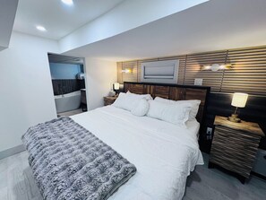 Una habitación de hotel moderna con una cama grande, una mesita de noche con lámpara, cabecera de madera, un banco empotrado y un baño visible en una esquina.