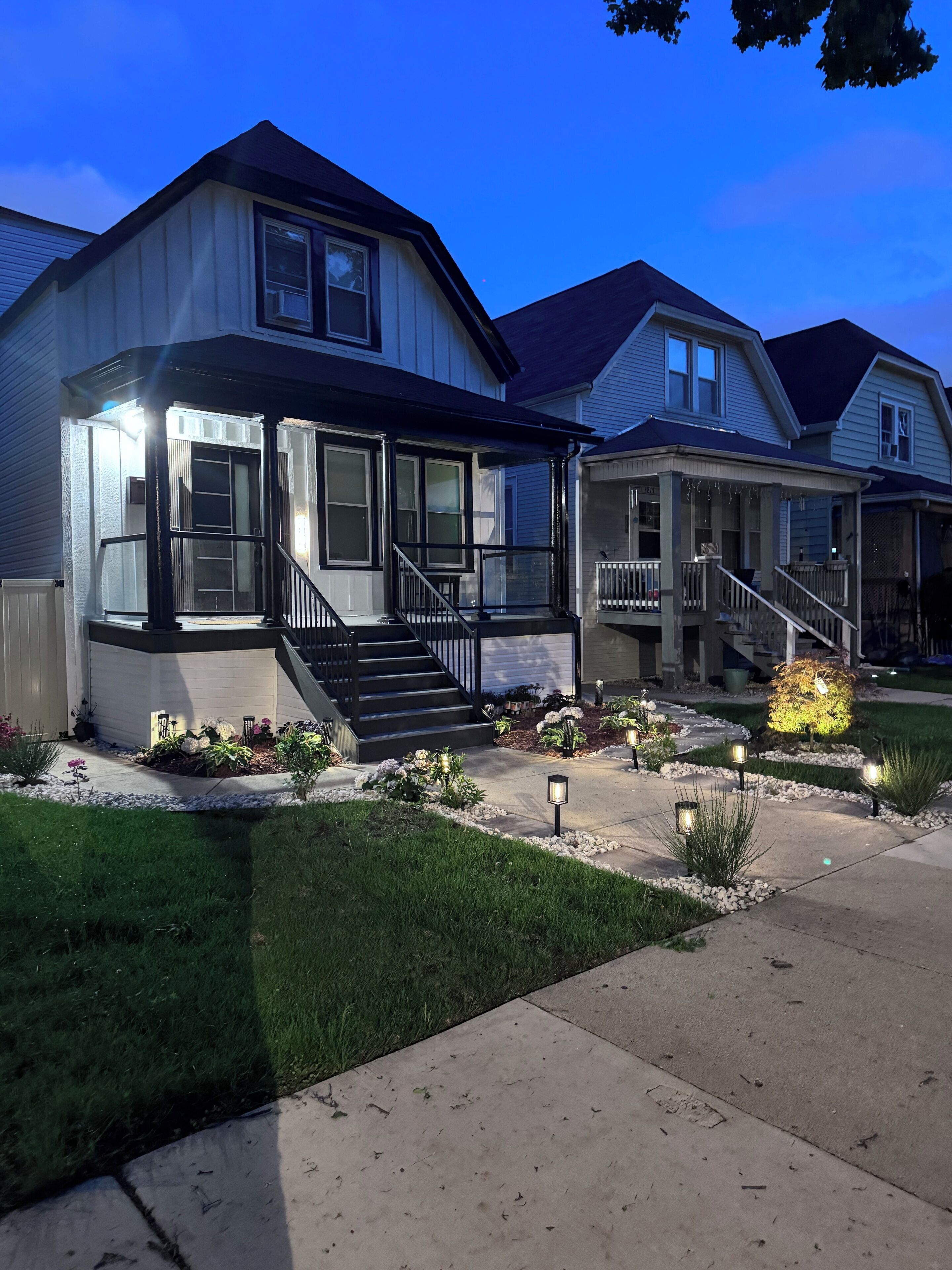 A two-story house with a covered porch, a front staircase, and a well-maintained front yard with plants and outdoor lighting.
