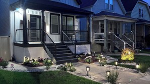 Ein zweigeschossiges Haus mit überdachter Veranda, einer Haustreppe und einem gepflegten Vorgarten mit Pflanzen und Beleuchtung.