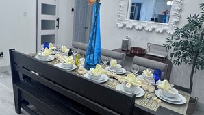 Ein Esstisch ist gedeckt mit weißen Tellern, Schüsseln und Gläsern, eine blaue Vase mit Blumen, eine Chandeliebre, ein Spiegel und eine Pflanze.