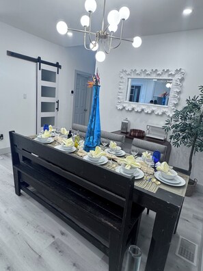 Una mesa de comedor dispuesta con platos, fuentes y vasos blancos, un jarrón azul con flores, una araña, un espejo y una planta.