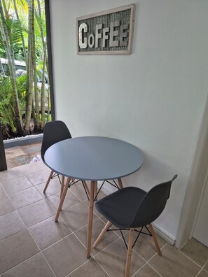 Ein runder Tisch mit zwei Stühlen, ein 'COFFEE' Schild an der Wand und Blick auf Palmen draußen.