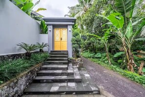 Uma trilha leva a um edifício com porta amarela, rodeado por vegetação exuberante.