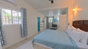 A bedroom with a bed, a window with curtains, a door, a bedside table, and a wall-mounted light fixture. - Family - Friendly 2BR Home - Safe Area, Walk to Food, Shops & Playing Field (Rodney Bay)