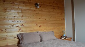 Un dormitorio con cabecera de madera, cama con ropa de cama estampada, mesita de noche con lámpara y una pequeña mesa con un florero y libros.