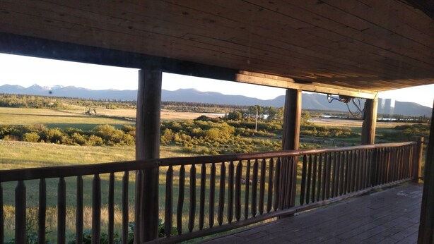 Una terraza de madera con barandilla, con vistas a un campo cubierto de hierba y montañas lejanas.