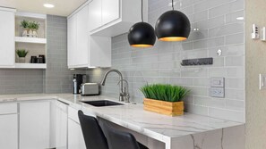 Eine moderne Küche mit weißen Schränken, einer grauen Fliesenrückwand, schwarzen Pendelleuchten, einem Holz-Pflanzgefäß mit Grünpflanzen und einer Marmorarbeitsplatte.