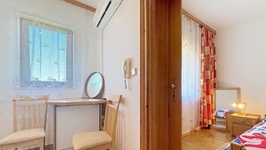 Ein Zimmer mit Schreibtisch aus Holz, zwei Stühlen, einem Heizkörper, einem Spiegel, einem Telefon, einem Bett mit gemustertem Überwurf und einem Fenster mit Spitzenvorhang.