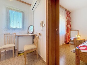Ein Zimmer mit Schreibtisch aus Holz, zwei Stühlen, einem Heizkörper, einem Spiegel, einem Telefon, einem Bett mit gemustertem Überwurf und einem Fenster mit Spitzenvorhang.