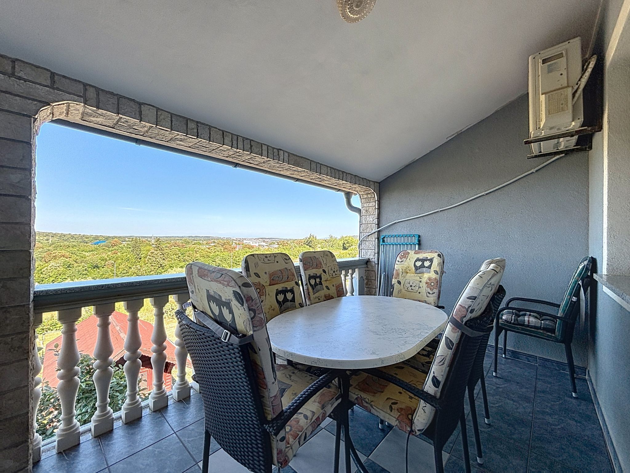 A balcony with a table and chairs, a view of greenery, and a wall-mounted air conditioning unit.