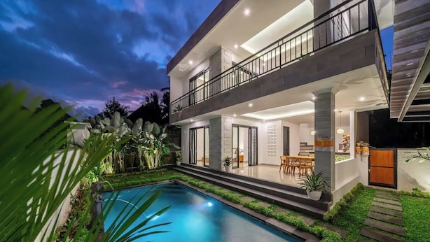 Una casa moderna de dos pisos con piscina, mobiliario exterior e iluminado balcón.