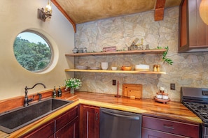 Una cocina con protector contra salpicaduras de piedra, repisas de madera, un ventanal redondo y un lavavajillas integrado.