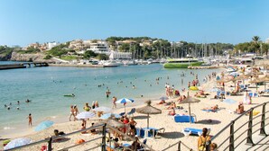 Una playa con numerosas tumbonas y sombrillas, un puerto deportivo con barcos, y un pueblo al fondo.