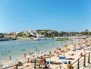 Ein Strand mit vielen Liegestühlen und Sonnenschirmen, eine Marina mit Booten und eine Stadt im Hintergrund.