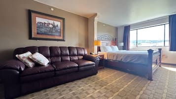 A hotel room with a dark brown leather sofa, a bed with a wooden headboard, a nightstand, a lamp, and a large window with curtains.