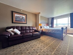 A hotel room with a dark brown leather sofa, a bed with a wooden headboard, a nightstand, a lamp, and a large window with curtains.