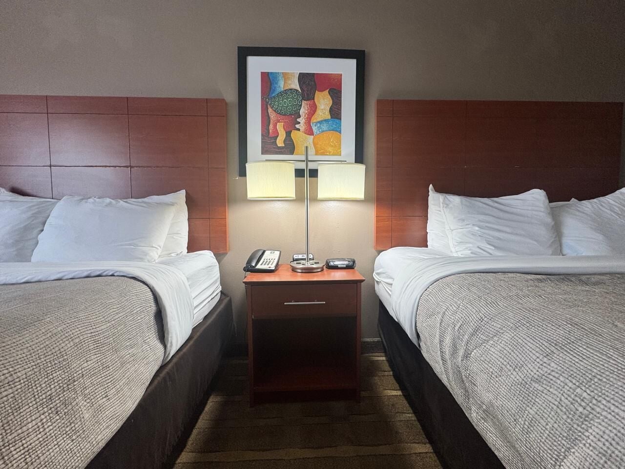 A hotel room with two beds, a nightstand with a lamp, a phone, and a framed artwork on the wall.