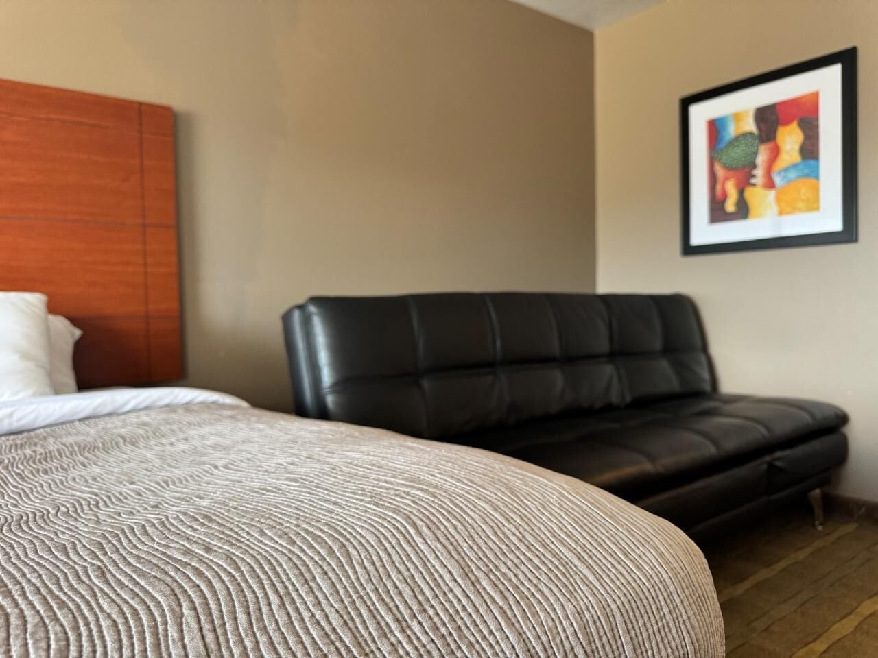 A hotel room with a bed, a black leather sofa, a wooden headboard, and a framed artwork on the wall.